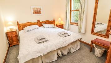 Een netjes opgemaakt bed met een houten hoofdeinde, twee bedlampjes, een nachtkastje met lamp, een raam met gordijnen, een spiegel, een klein bankje en een aan de muur hangend schilderij.