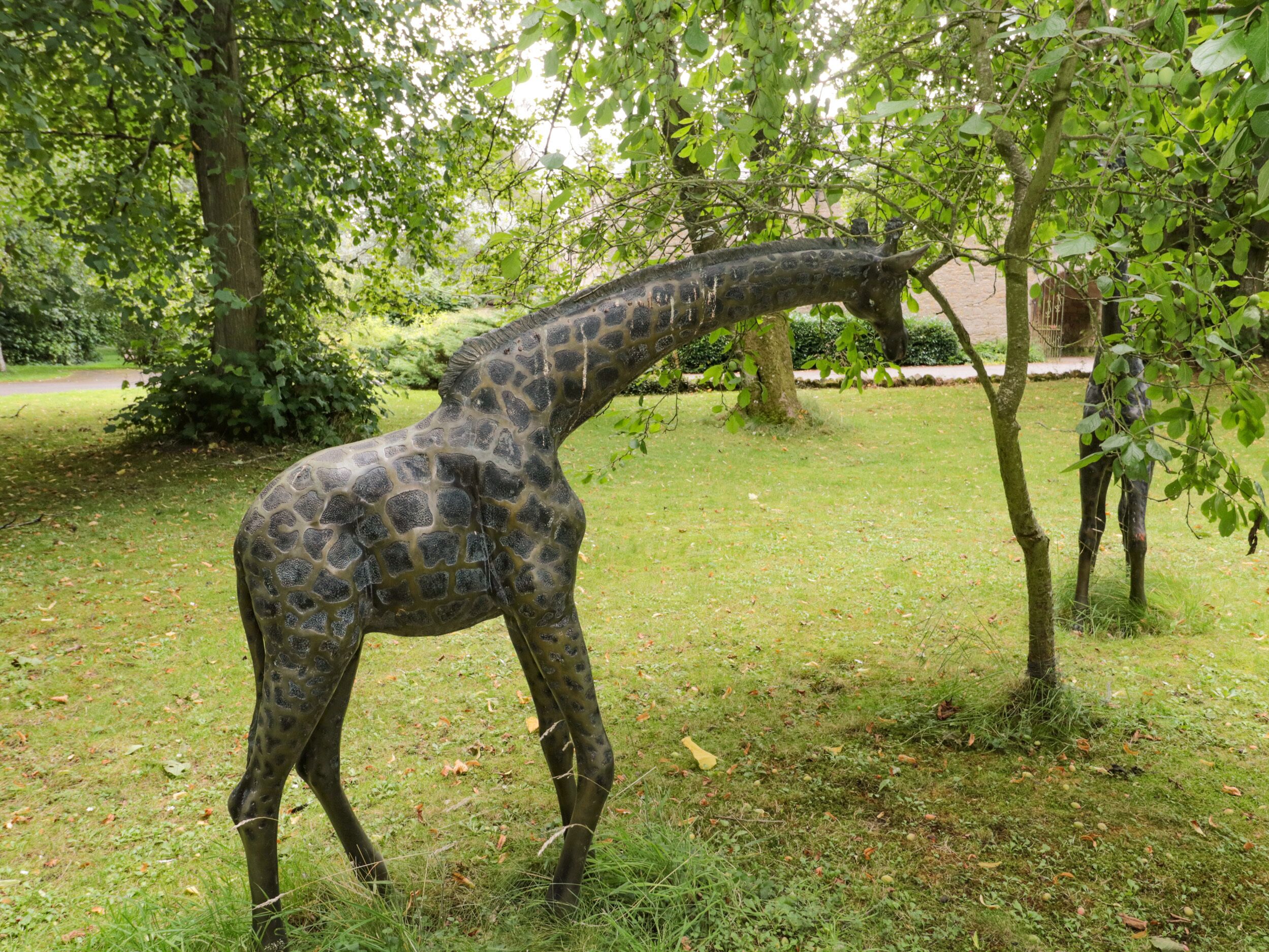 Eine Giraffenskulptur in einer grünen Umgebung mit Bäumen und einer Backsteinmauer im Hintergrund.