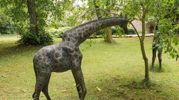 Eine Giraffenskulptur in einer grünen Umgebung mit Bäumen und einer Backsteinmauer im Hintergrund.