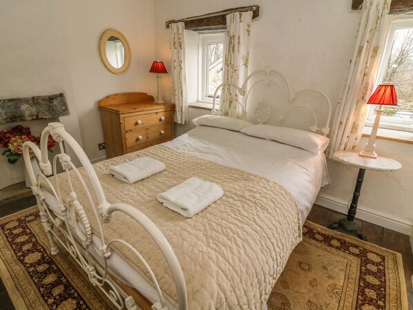 A bedroom with a bed, a wooden dresser, a round table with a lamp, and a mirror on the wall. - Beckside Cottage (Carnforth)