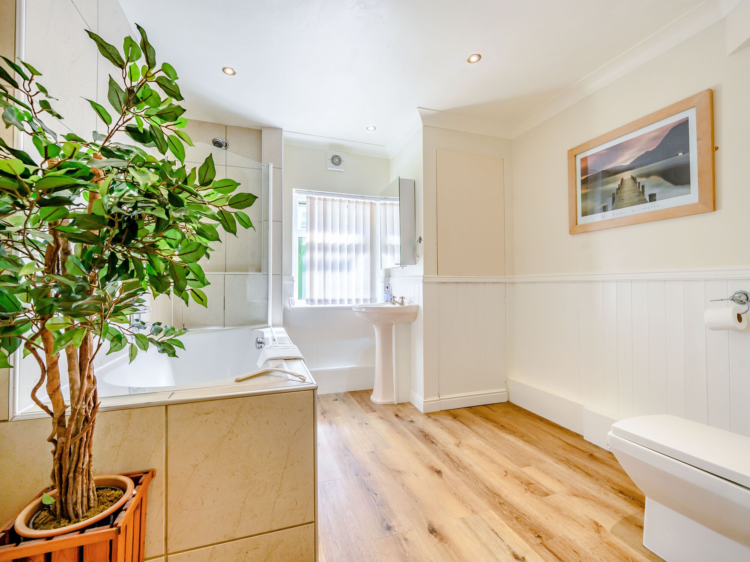 A modern bathroom with a wooden floor, a white bathtub, a sink, and a toilet. There is a potted plant and a framed picture on the wall.