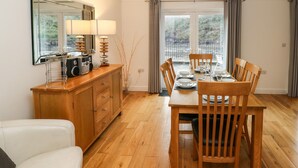 A dining area with a wooden table set for four, a wooden cabinet, a white sofa, and a large window with curtains. - Curlew (Milford Haven)