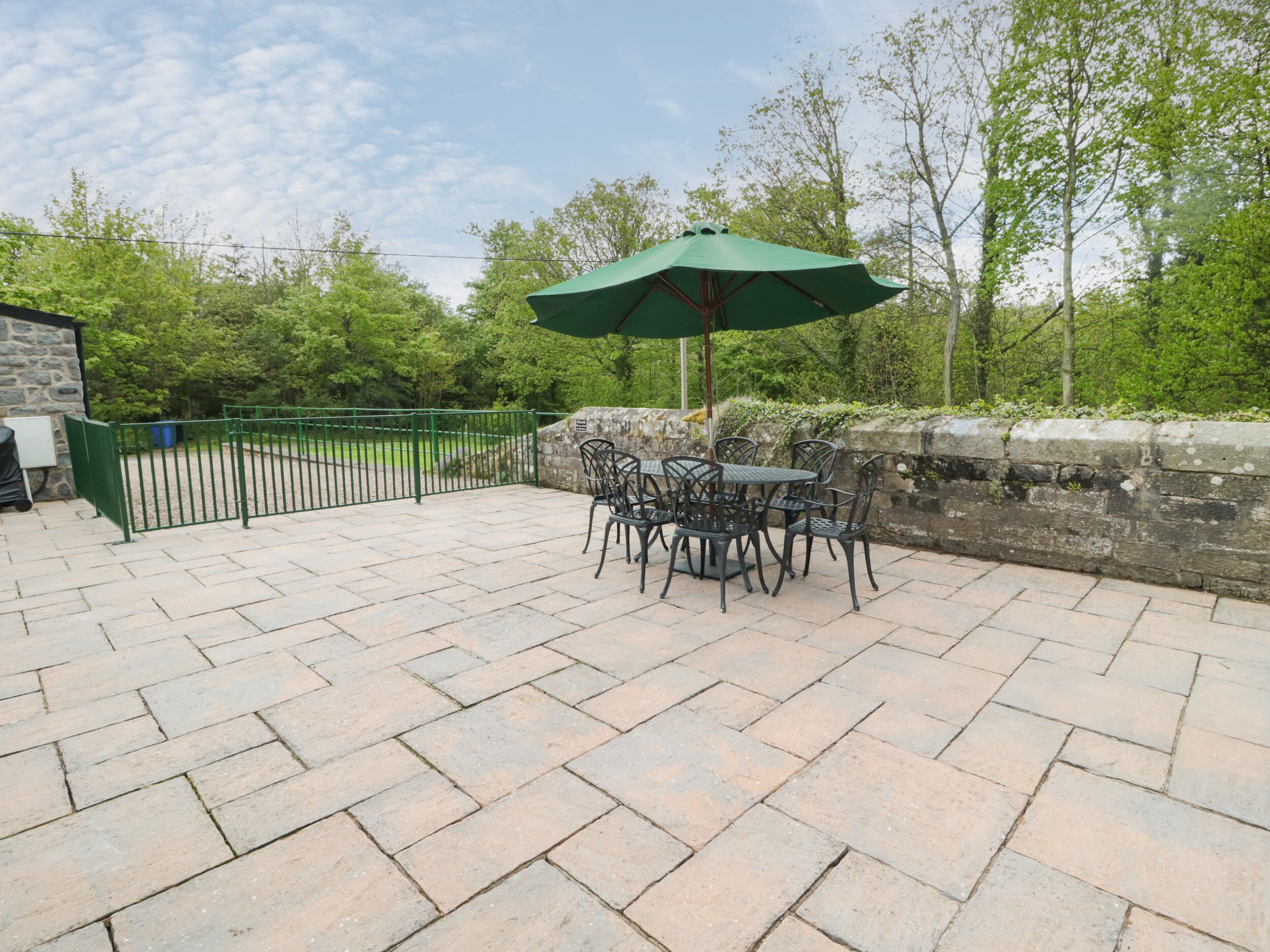 An outdoor patio with stone flooring, a green umbrella, and metal chairs.