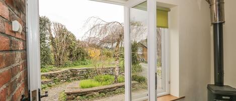 A room with a stone wall, a wooden door, a window with a view of a garden, and a wood-burning stove.