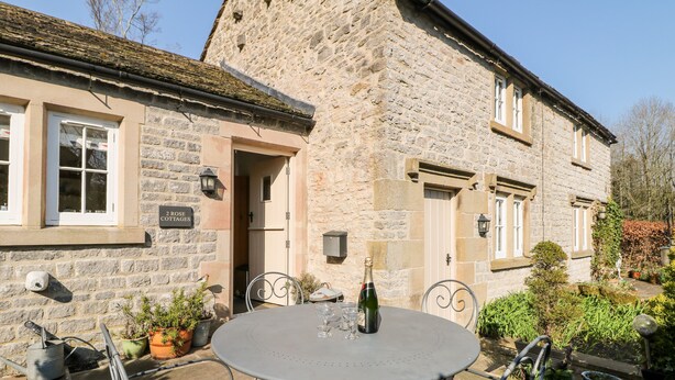 Una cabaña de piedra con una mesa redonda y sillas de metal en el exterior, una botella de champán y un letrero que dice "Rose Cottages".