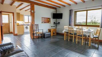 A spacious living area with a dining table set for four, a television mounted on the wall, and a large window offering a view of the outdoors.