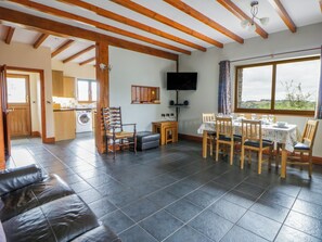 A spacious living area with a dining table set for four, a television mounted on the wall, and a large window offering a view of the outdoors. - Y Bwthyn (Tregaron)