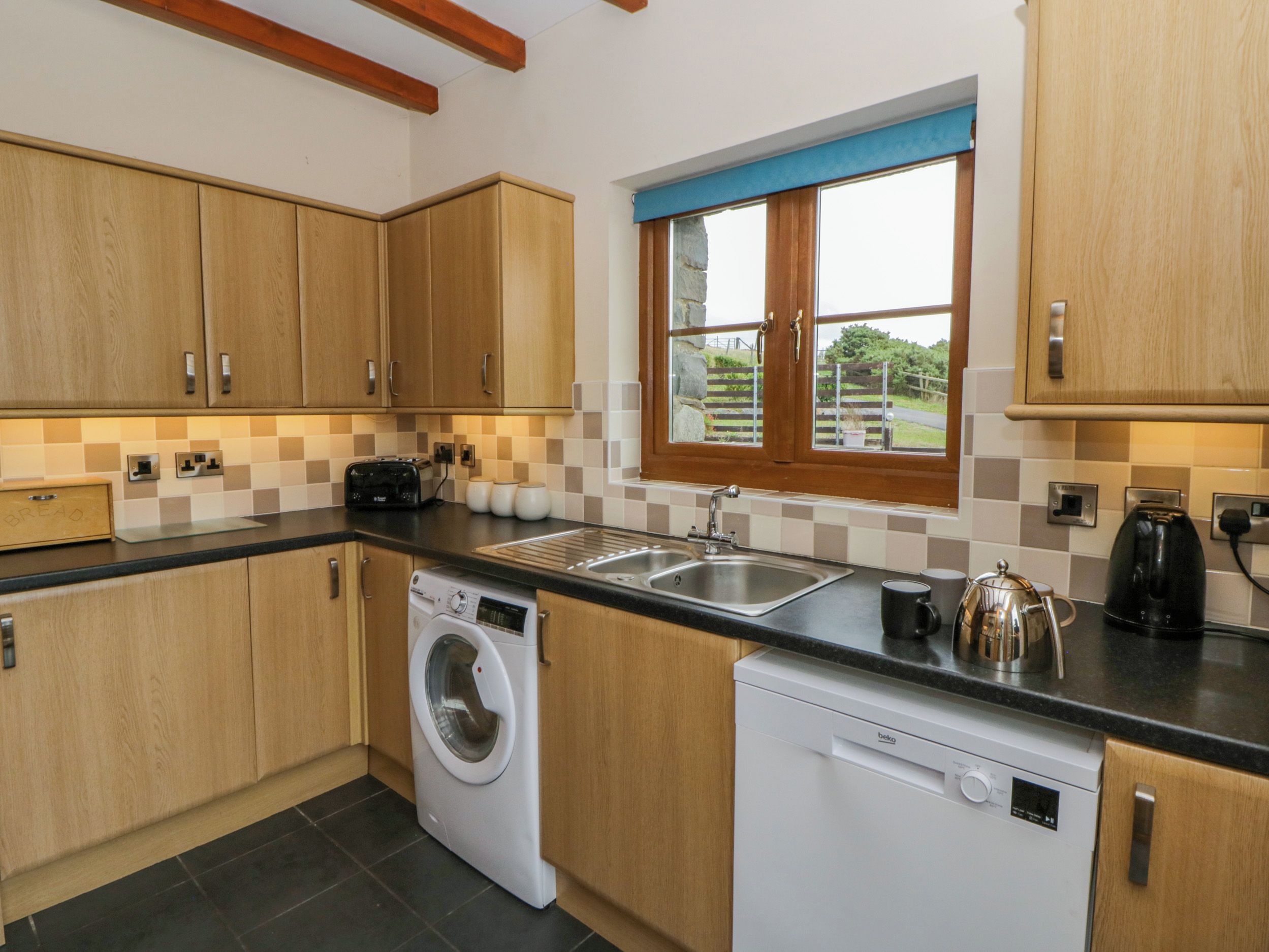 A compact kitchen with wooden cabinets, a built-in washing machine, a dishwasher, a window with a view, and a tiled backsplash.