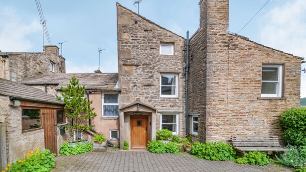 A stone building with a wooden door, a small garden, and a paved courtyard. - Mill Cottage (Hawes)