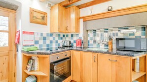 A kitchen with wooden cabinets, a microwave, a kettle, and a tiled backsplash. - Mill Cottage (Hawes)