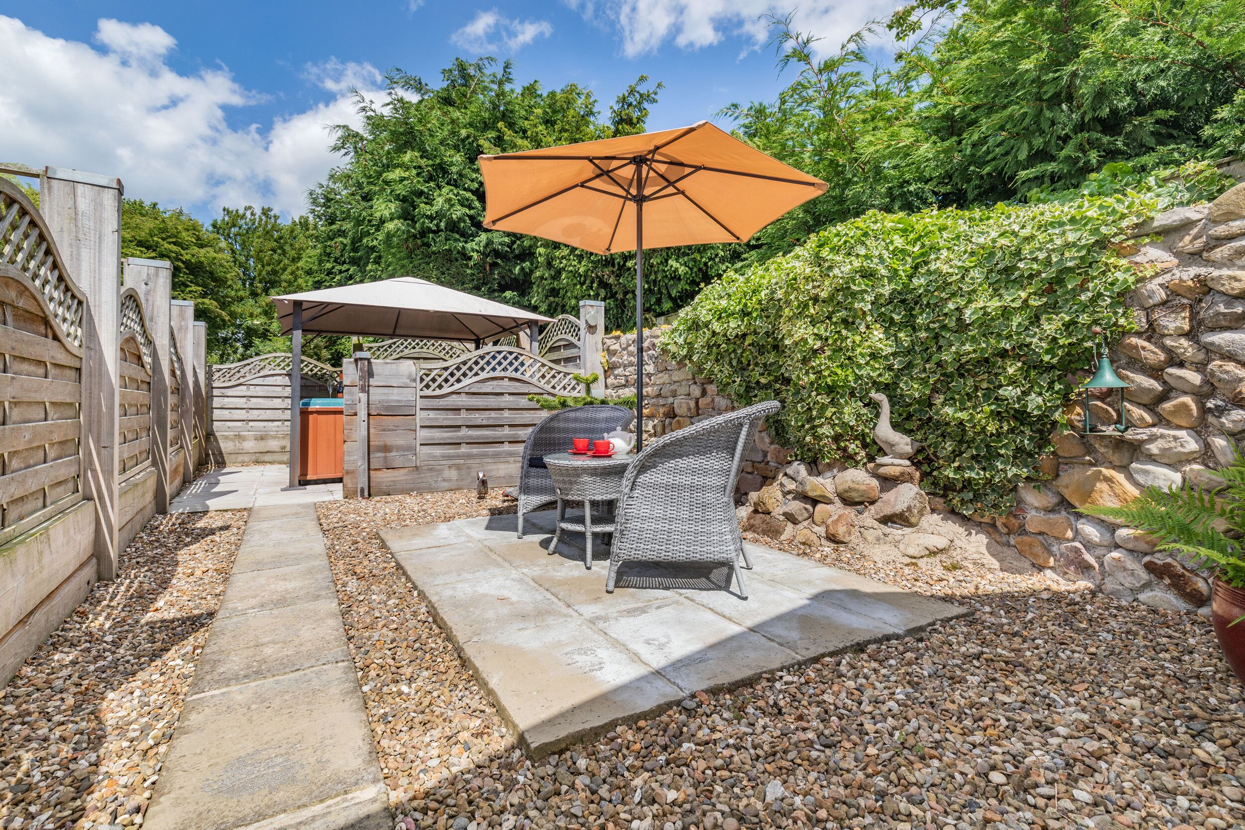 An outdoor patio area with a stone pathway, a table with chairs, a barbecue grill, and an umbrella.