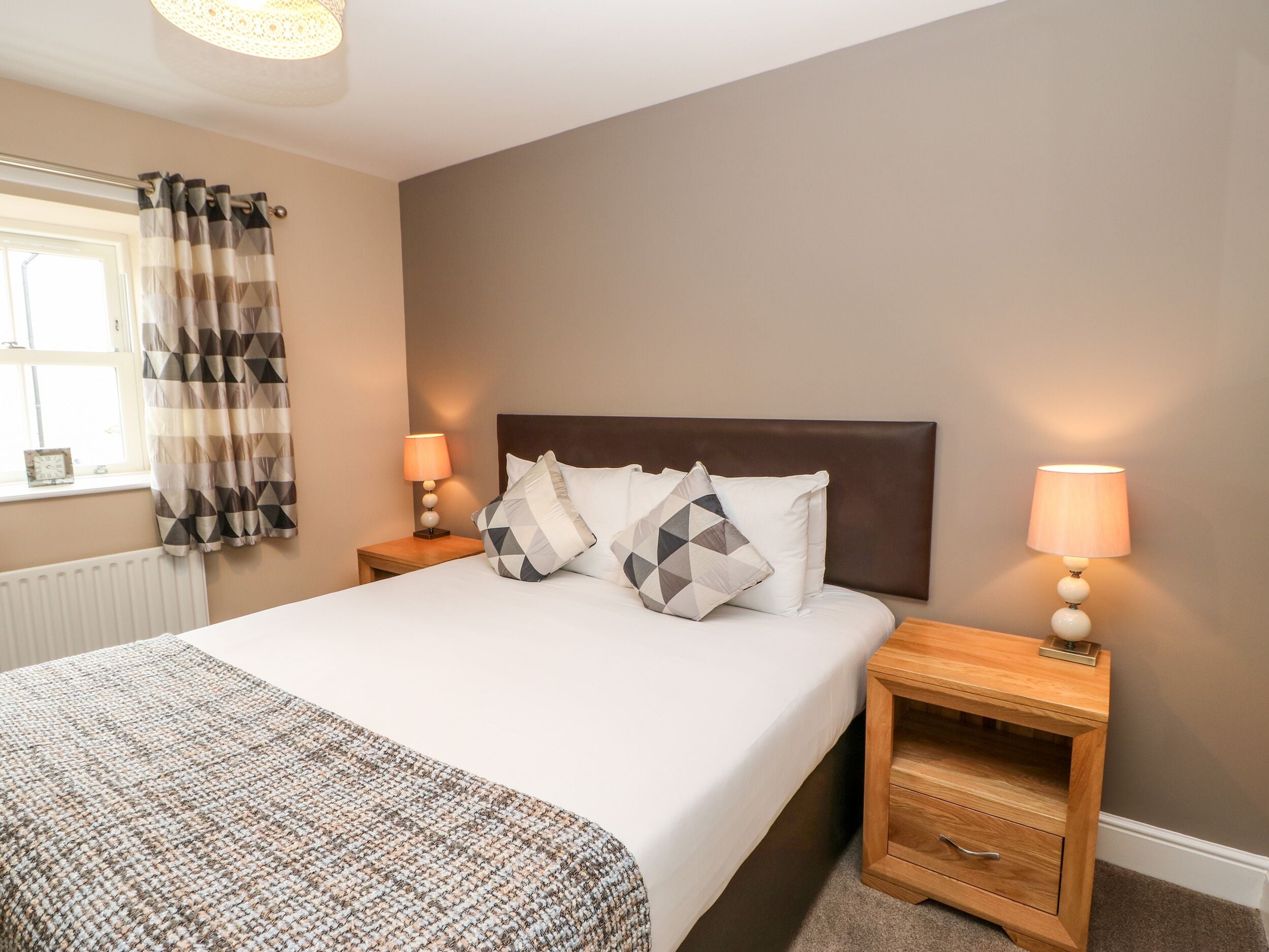 A neatly made bed with a headboard, two bedside tables with lamps, a window with curtains, and a radiator.