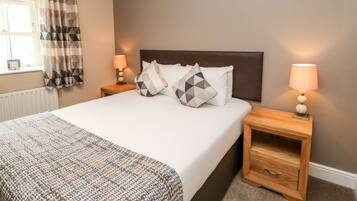 A neatly made bed with a headboard, two bedside tables with lamps, a window with curtains, and a radiator.