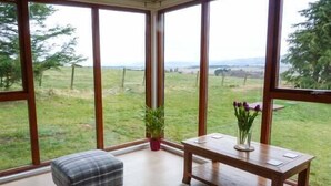 A room with large windows, a plaid ottoman, a small table with a vase of flowers, and a grey rug. - Pine Lodge (Dingwall)