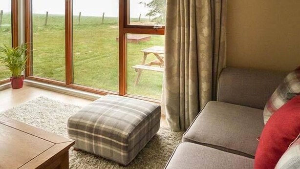A living room with a large window, a wooden table, a plaid ottoman, and a sofa. - Pine Lodge (Dingwall)