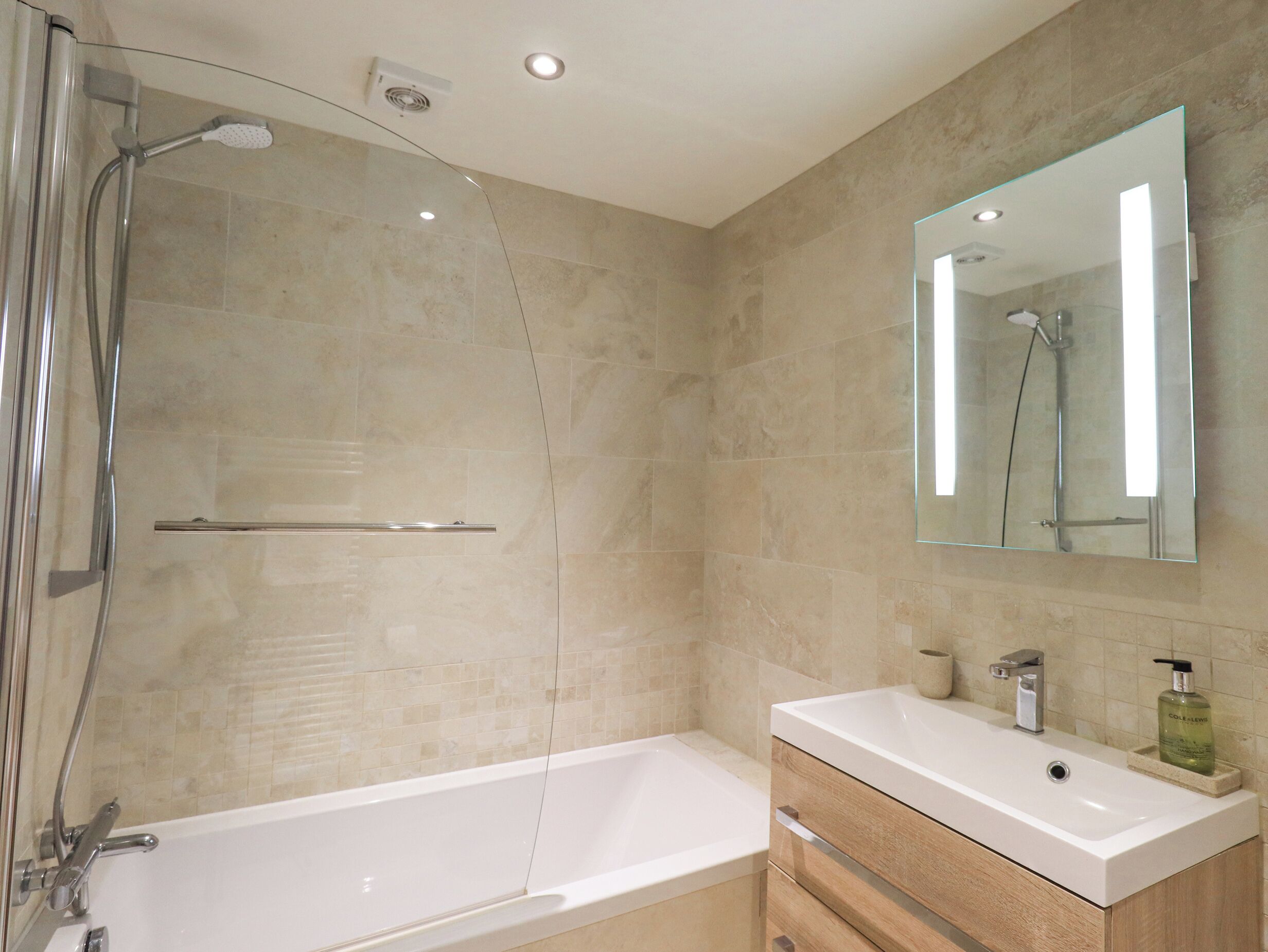 A modern bathroom with a glass shower enclosure, a white bathtub, a vanity with a mirror, and a beige tiled wall.
