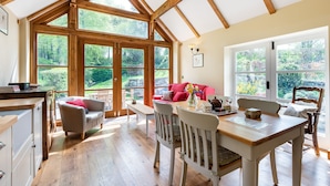 A spacious room with a large wooden-framed window, a dining table with chairs, a sofa, and a kitchen area.