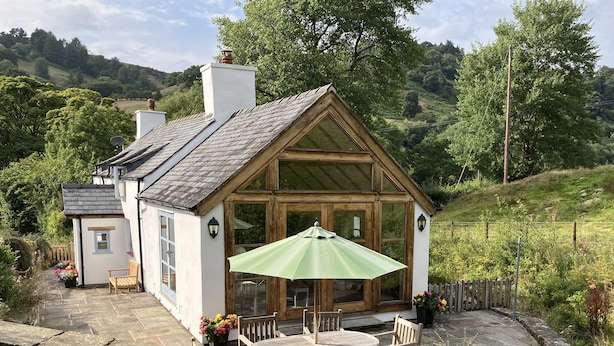 A cozy cottage with a stone patio, wooden furniture, and a green umbrella.