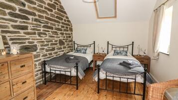 A bedroom with two beds, a wooden dresser, a stone wall, and a window with curtains.