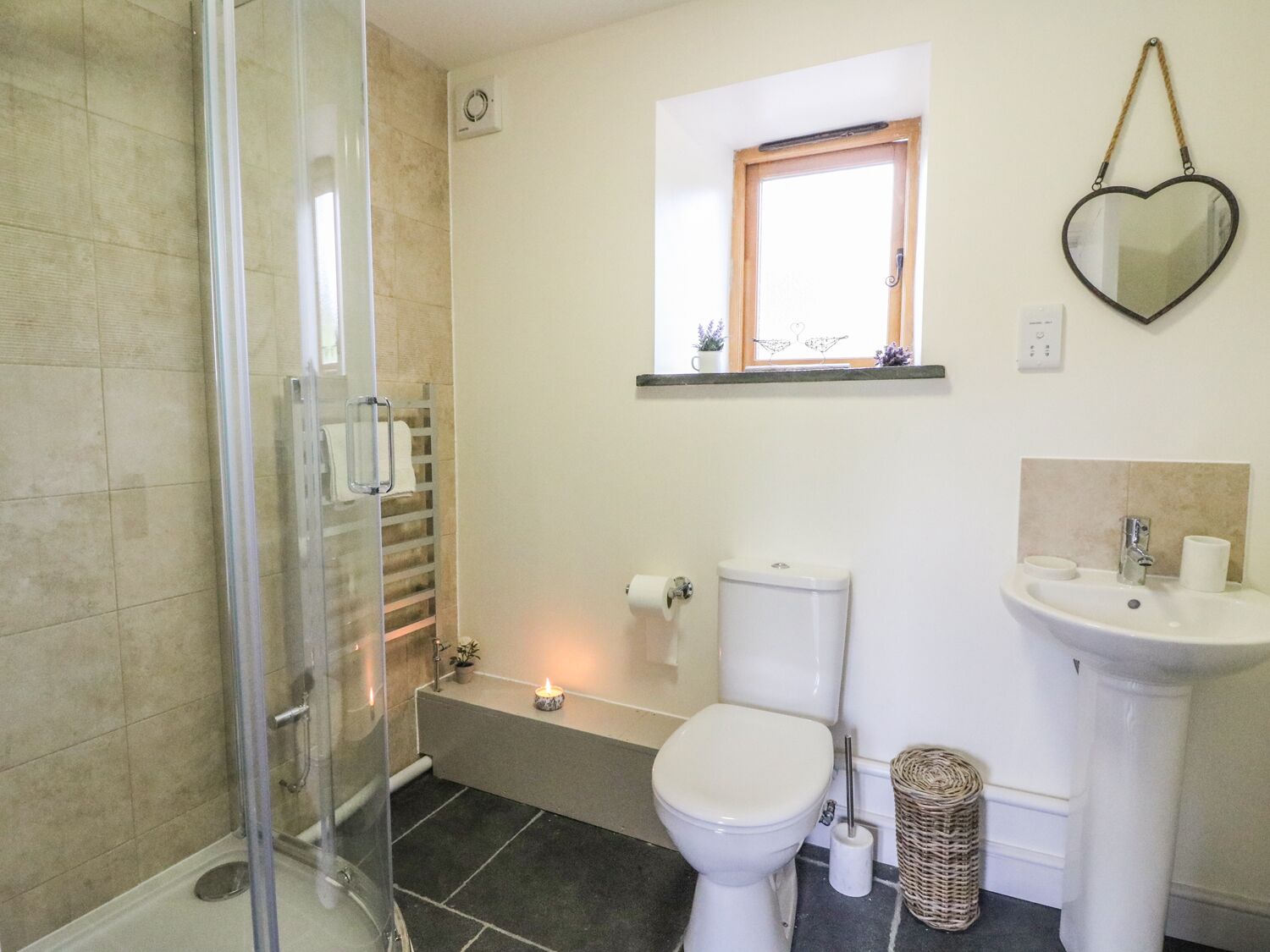 Baño con cabina de ducha de vidrio, inodoro, lavamanos y una ventana pequeña con un espejo decorativo con forma de corazón.