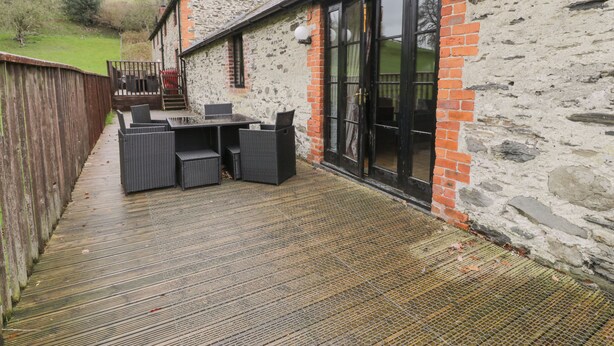 An outdoor patio with a table and chairs, a wooden deck, and a stone building with a glass door. - The Dairy (Corwen)