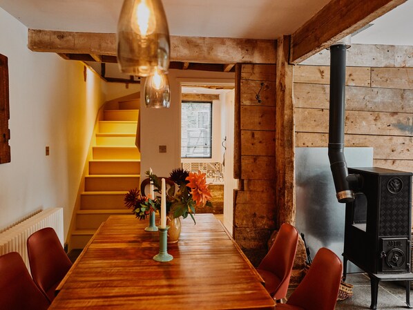 A wooden dining table with chairs, a vase with flowers, and a candle. There is a staircase leading to a window with blinds. A black wood stove is visible in the corner. - The Hayloft (Ross-on-Wye)