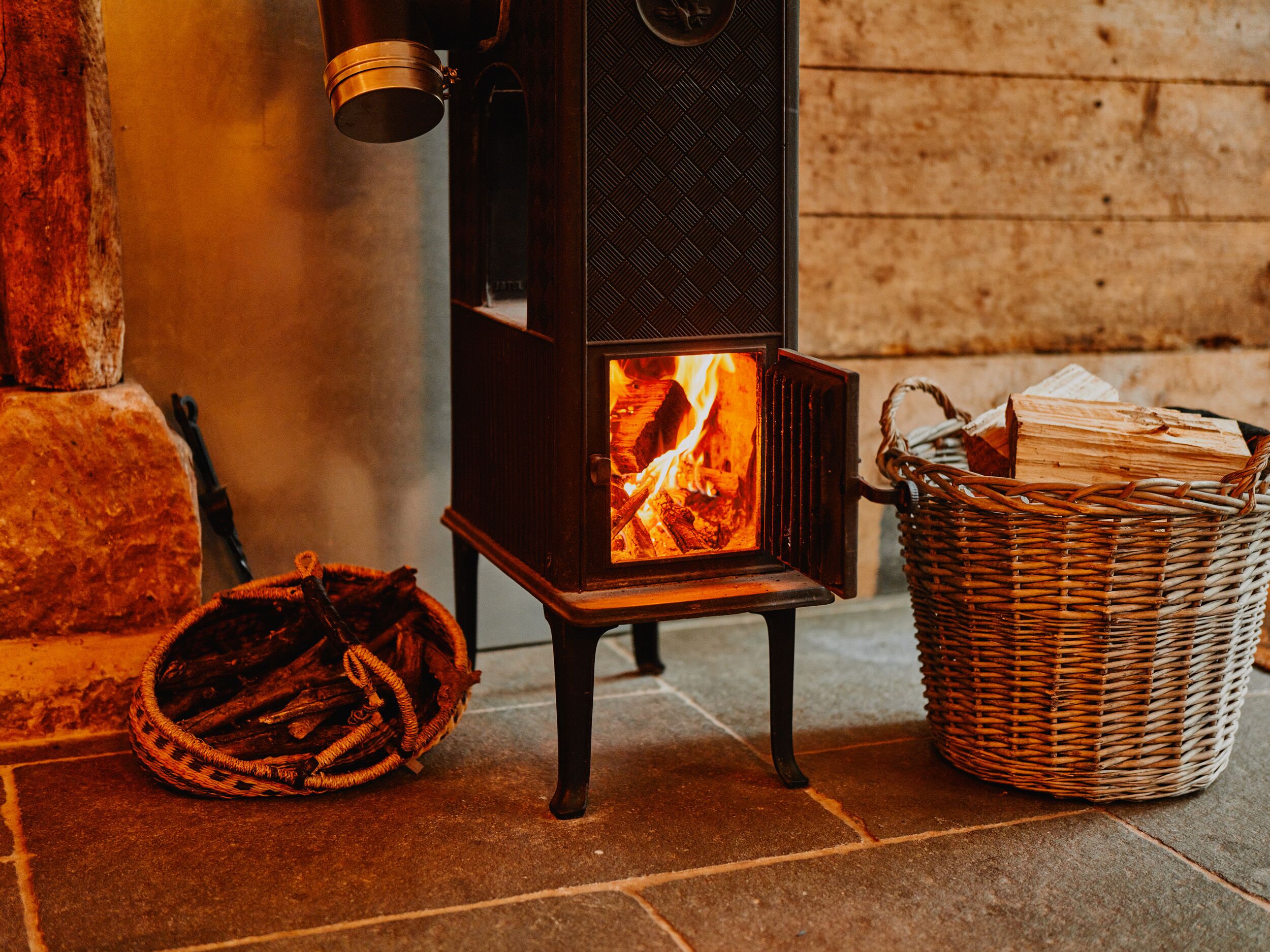 A wood-burning stove with logs, a basket of firewood, and a stone wall in the background.