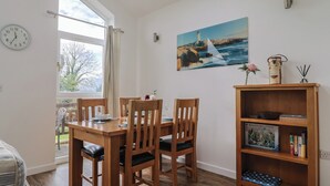 Un coin repas avec une table et des chaises en bois, un tableau représentant un phare accroché au mur, une horloge et une bibliothèque remplie de livres et d’un jeu de puzzle.