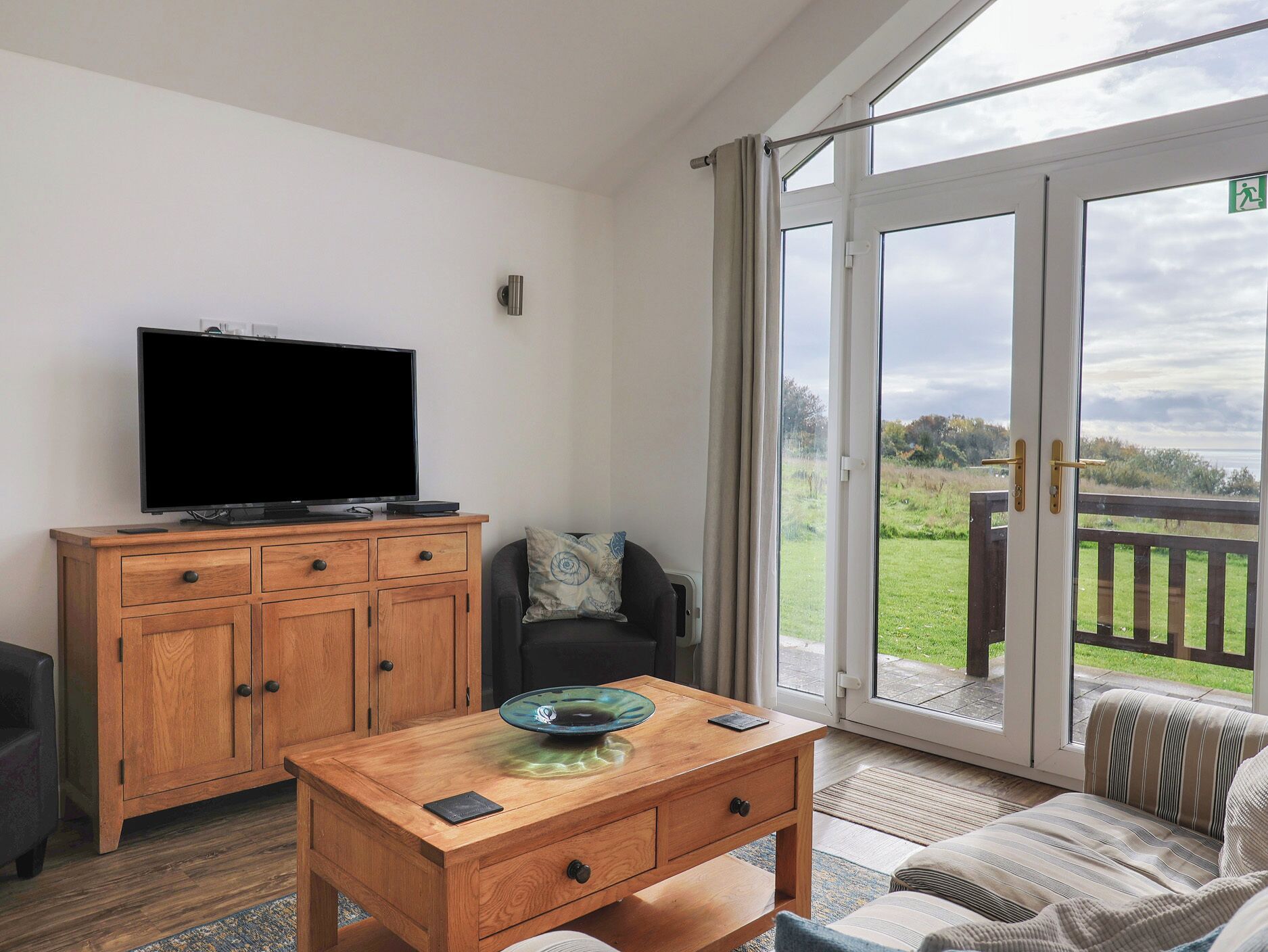 Una habitación con un armario de madera, un televisor, una mesa de centro con un cuenco decorativo, un sofá y un ventanal corredero que da a una terraza exterior.