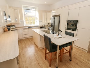 A modern kitchen with white cabinetry, a central island with seating, a built-in oven, and a coffee machine. - Hideaway House at The Hideaway (Tenby)