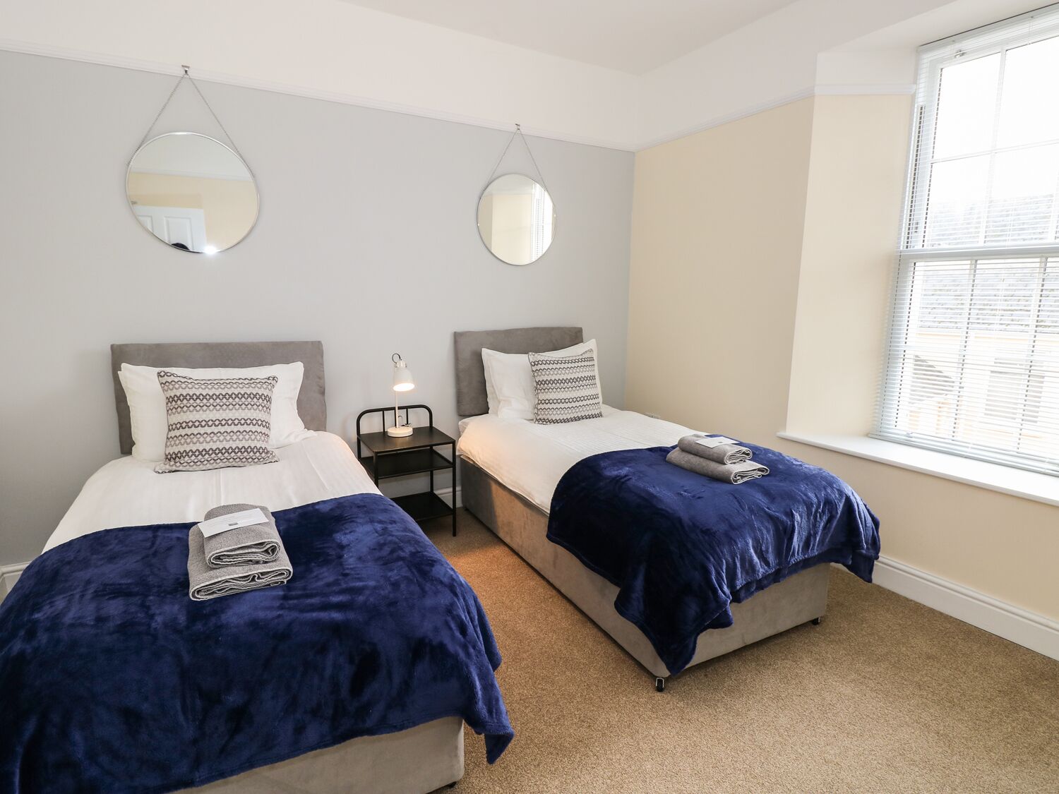A twin bedroom with two beds, a nightstand, a lamp, and a window with blinds.