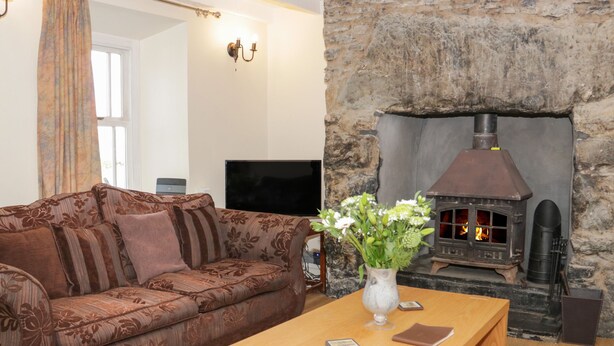 A living room with a stone fireplace, a brown patterned sofa, a wooden coffee table, and a vase with flowers. - Bryn Dedwydd Farmhouse (Betws-Y-Coed)