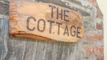A rustic wooden sign with the words "THE COTTAGE" carved into it, mounted on a weathered brick wall.
