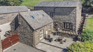 A stone building with a sloped roof, a gated entrance, and a patio area with outdoor seating. - Thimble Cottage (Ulverston)