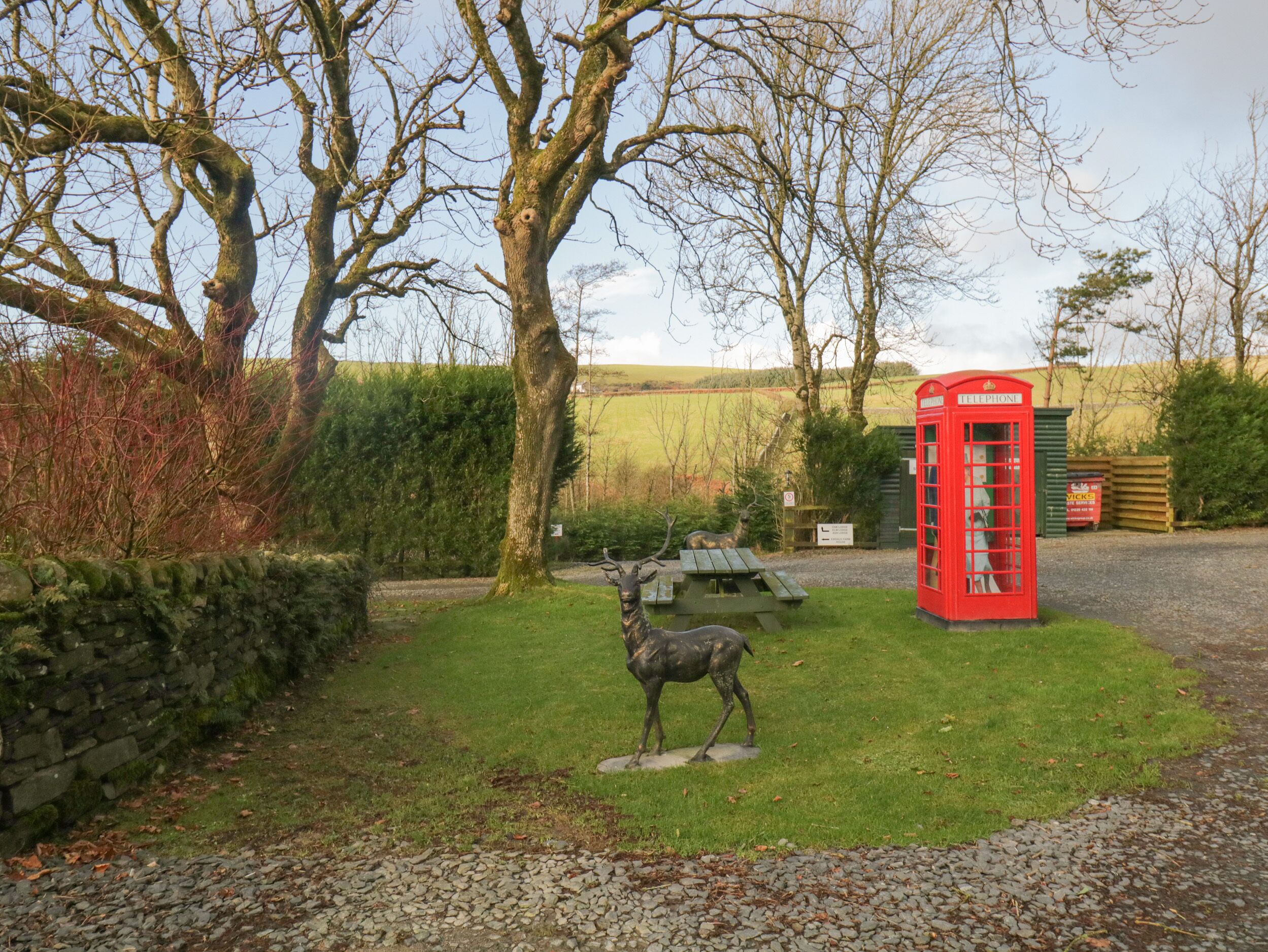 A statue of a deer in a grassy area with a red telephone booth and a picnic table in the background.