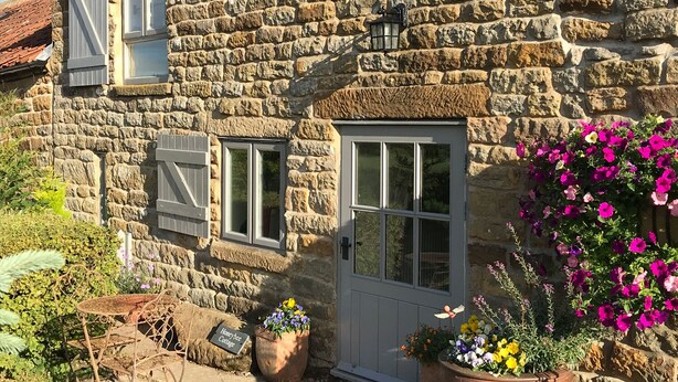 Une petite maison en pierre avec une porte blanche, deux fenêtres avec volets et un petit jardin agrémenté de plantes en pot et de fleurs.