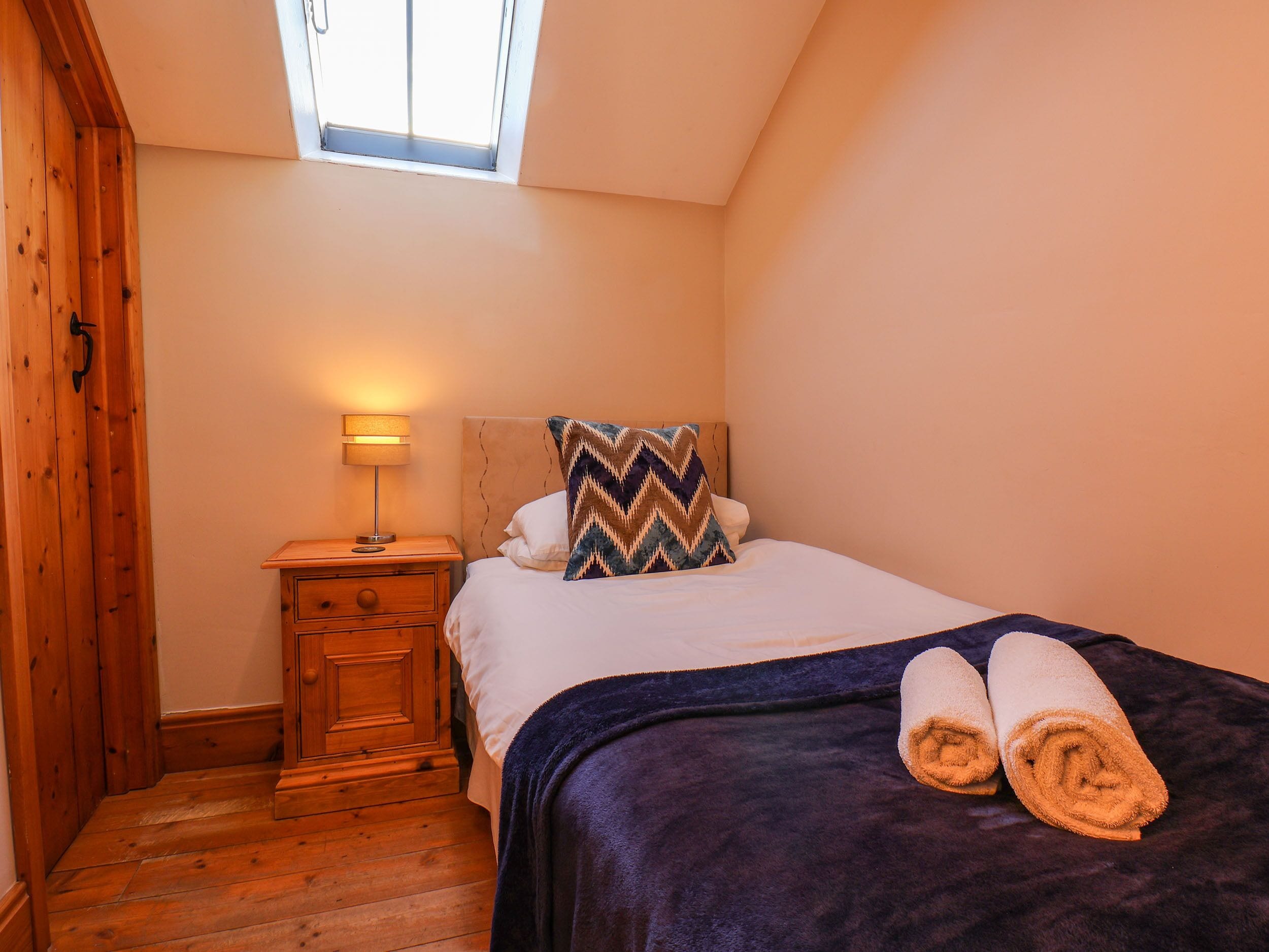 Un dormitorio con piso de madera, una cama con manta azul oscuro, dos toallas enrolladas, una mesita de noche con lámpara y una ventana con luz natural.