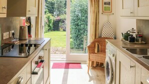 Een keuken met een ingebouwde wasmachine, een eethoek met een rieten stoel, en uitzicht op een tuin door een glazen deur.