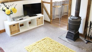 A cozy living room with a TV, white entertainment unit, yellow rug, and a wood-burning stove. - The Hayloft (Thetford)