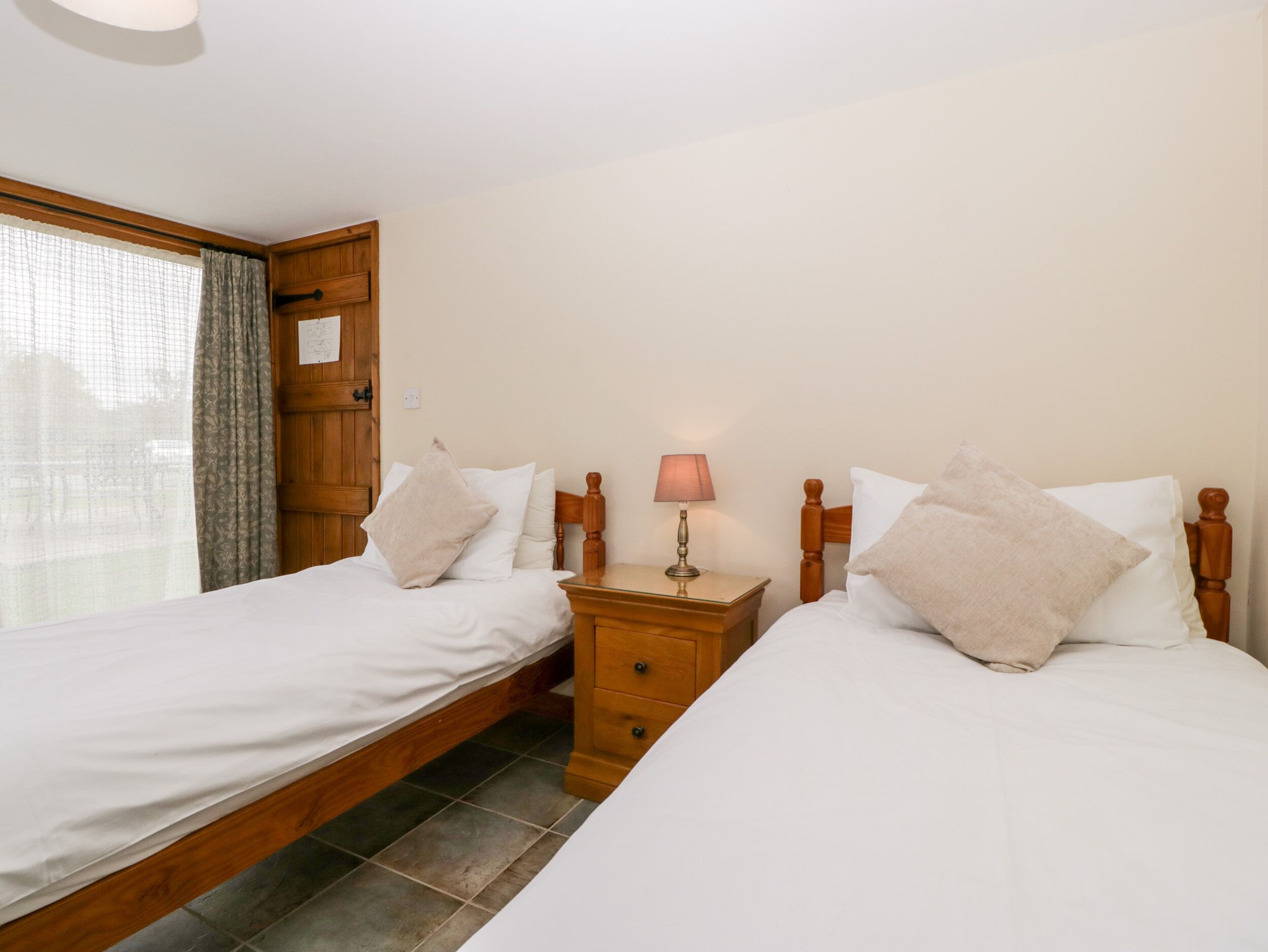 A bedroom with two single beds, a wooden nightstand with a lamp, and a wooden door with a window.