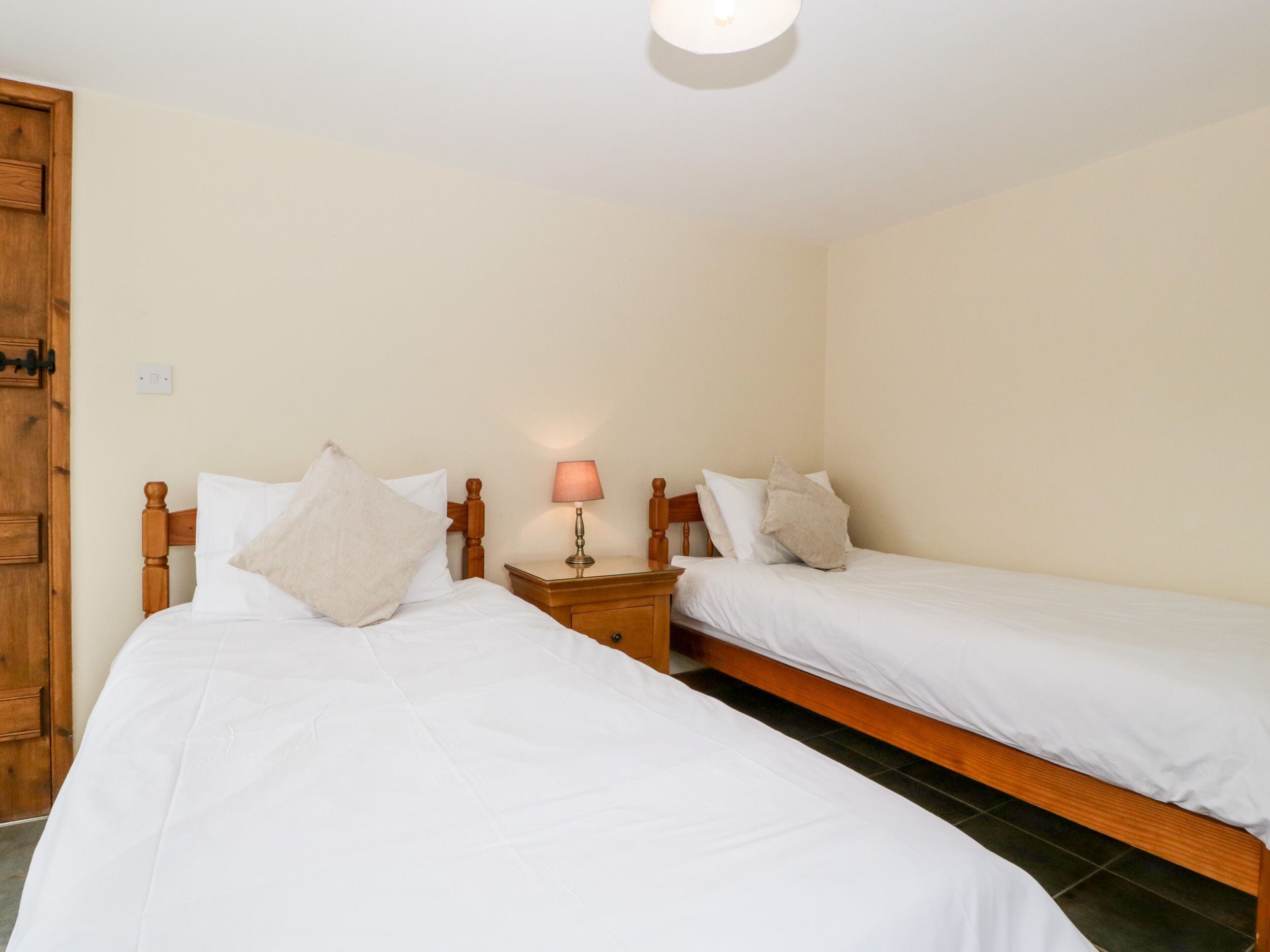 A room with two single beds, wooden headboards, a bedside table with a lamp, and a wooden door.