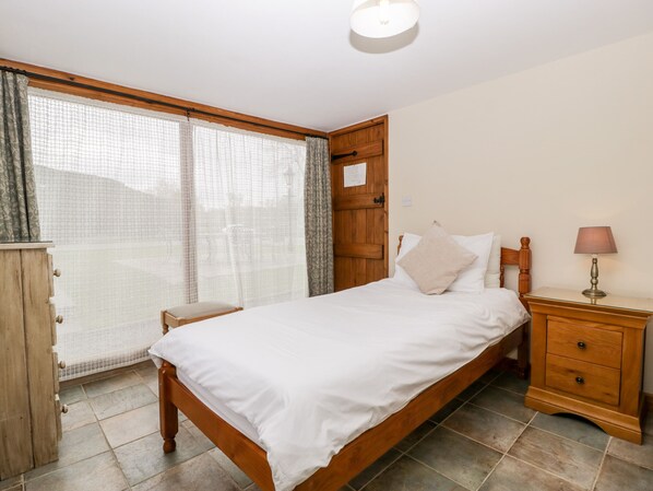 Un dormitorio con una cama de madera, una mesita de noche con lámpara, un armario de madera y una ventana con cortinas translúcidas.