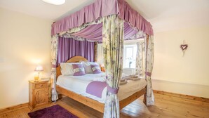 A four-poster canopy bed with a wooden frame and purple drapes, a wooden nightstand with a lamp, and a purple rug on a wooden floor. - Ty Nant (Llanrwst)