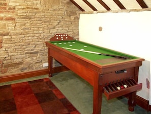 Una mesa de billar con estructura de madera y fieltro verde, frente a una pared de piedra y una alfombra roja.