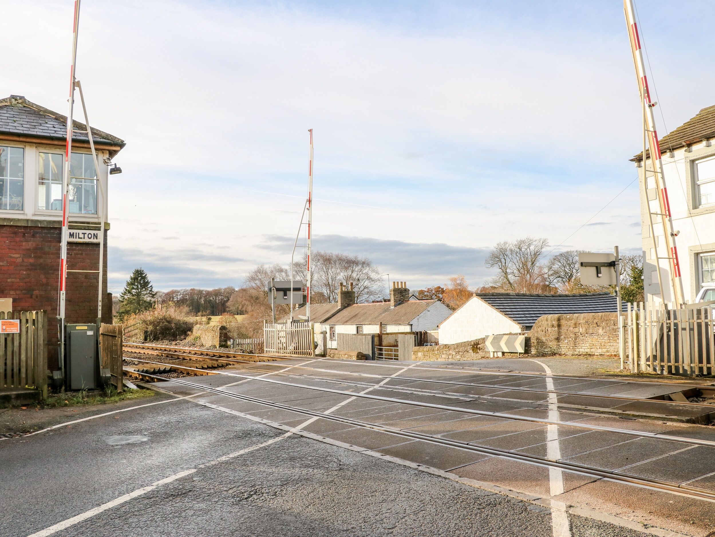 A railway crossing with a barrier gate, a signal box labeled 'MILTON', and a stone wall.