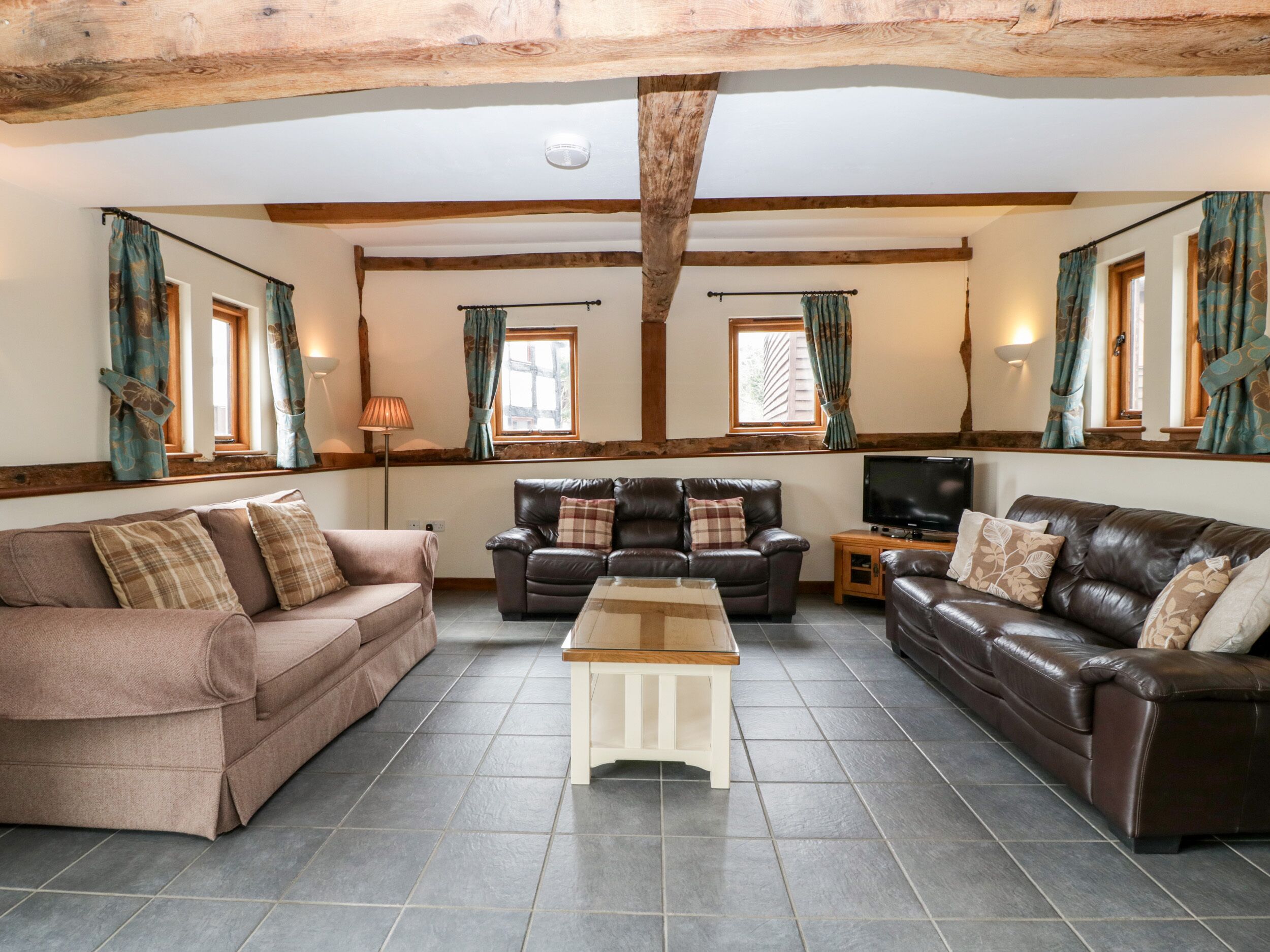 A living room with two sofas, a coffee table, and a TV. The room has wooden beams on the ceiling and large windows with curtains.