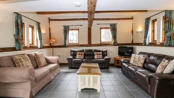 A living room with two sofas, a coffee table, and a TV. The room has wooden beams on the ceiling and large windows with curtains.