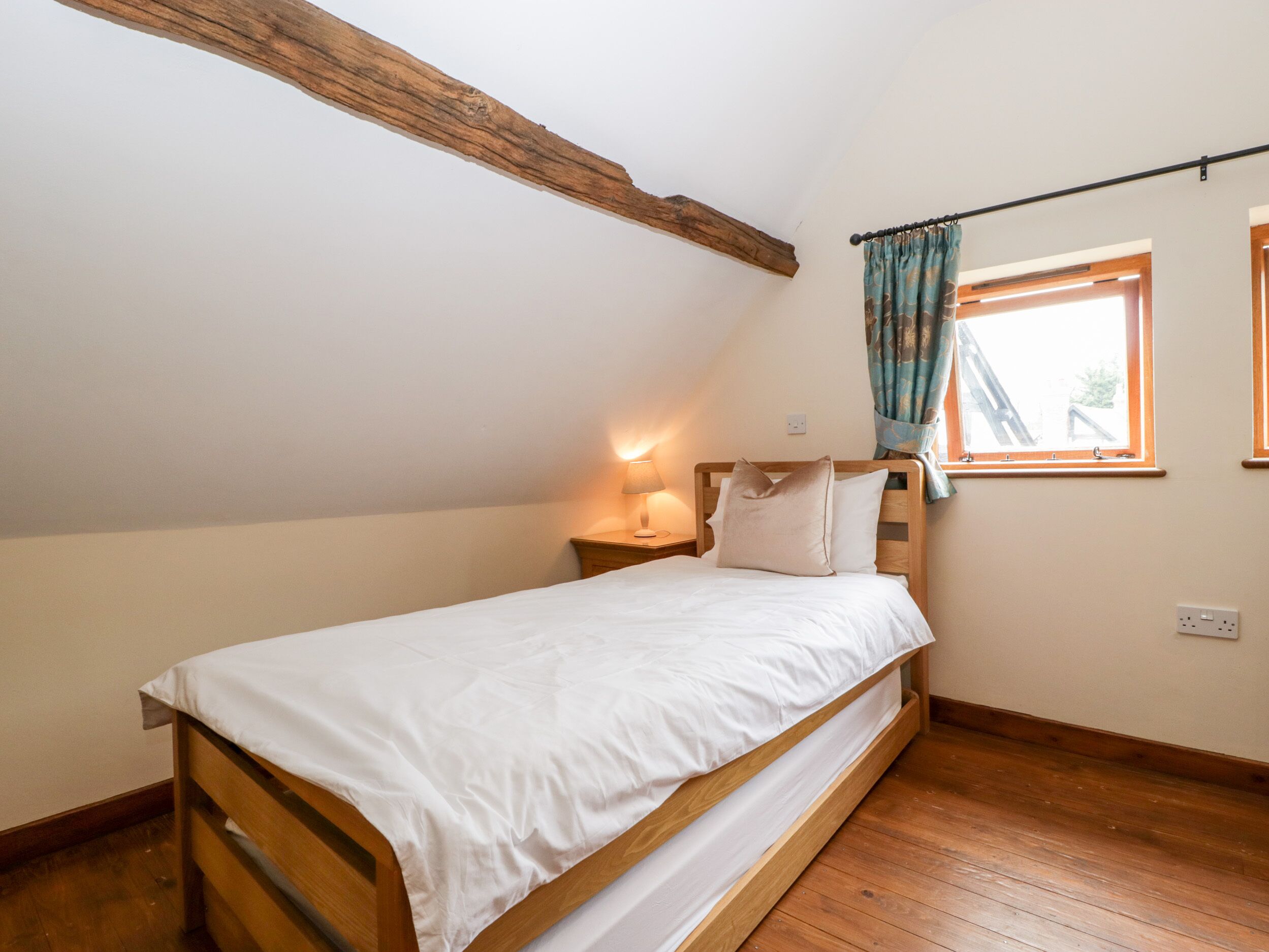 Een kleine kamer met een enkel bed, schuine balkenplafond, een raam met gordijnen en een bedlamp.