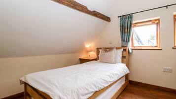 Een kleine kamer met een enkel bed, schuine balkenplafond, een raam met gordijnen en een bedlamp.