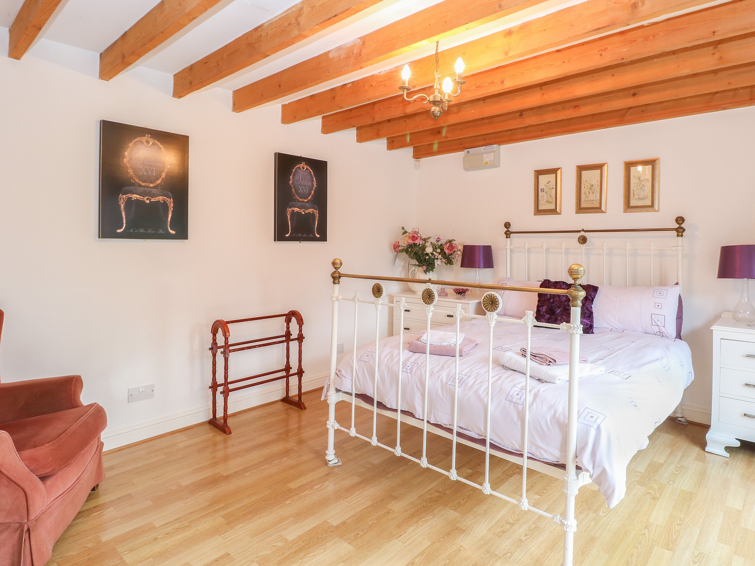 A bedroom with a white metal bed frame, wooden beams on the ceiling, a wooden chair, and a pink sofa.